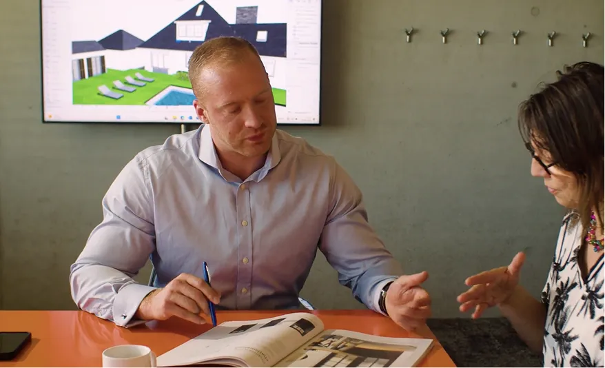 Ein Mann und eine Frau diskutieren über ein aufgeschlagenes Buch vor einem Bildschirm mit 3D-Hausmodell.