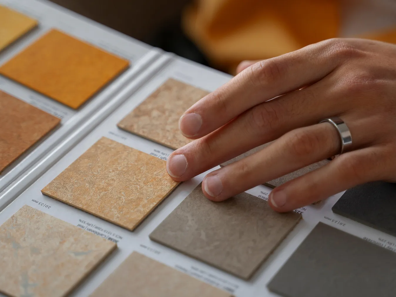 Hand mit Silberring betrachtet verschiedene Farb- und Materialproben von Fliesen oder Oberflächen.