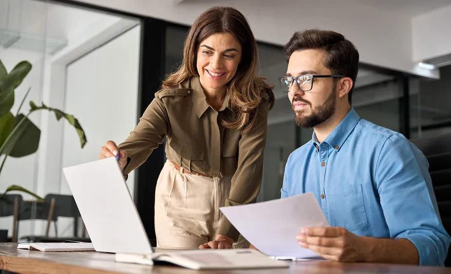 Zwei Kollegen besprechen Dokumente im modernen Büro, eine Frau zeigt auf Laptop-Bildschirm, ein Mann hält Papier.