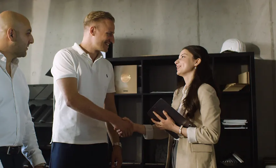 Drei Personen in einem Büro begrüßen sich, während zwei sich die Hand geben und lächeln.