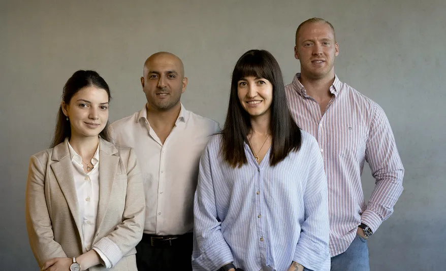 Vier lächelnde Erwachsene, zwei Frauen und zwei Männer, vor einer grauen Wand stehend.