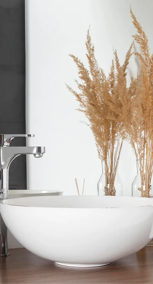 Weißes rundes Waschbecken mit verchromtem Wasserhahn vor einem Spiegel und getrockneten Gräsern in Glasvasen auf einer Holzablage.