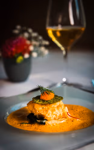 Middagstallerken med en fiskerett i kremet saus, pyntet med dill og rogn, med et vinglass og en blomsterpotte i bakgrunnen.