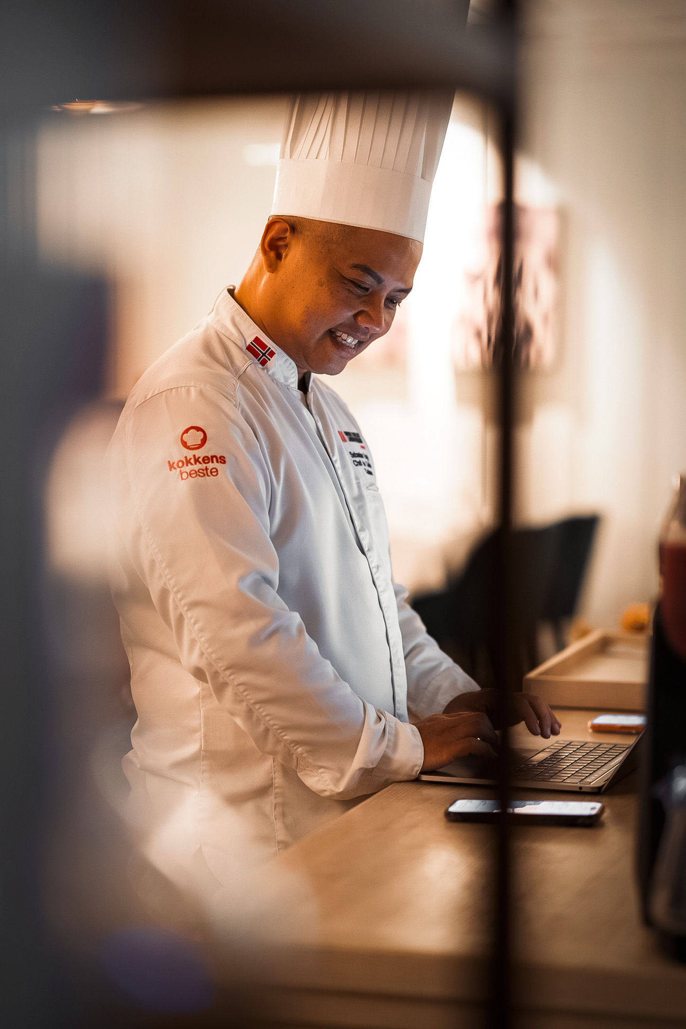 Kokk i hvit uniform med kokkelue, som smiler mens han bruker en bærbar datamaskin ved et trebord.