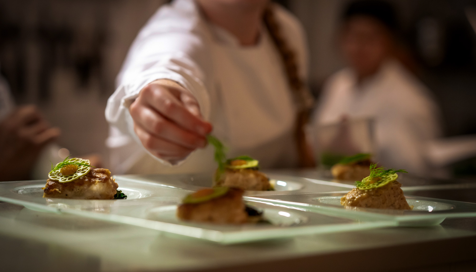 Kokk som pynter gourmet-retter på firkantede tallerkener i et profesjonelt kjøkken.