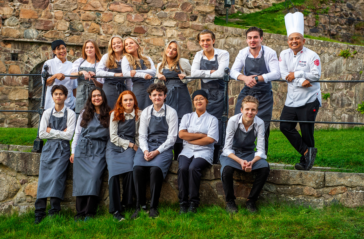 En gruppe unge kokker og to profesjonelle kokker poserer utendørs foran en steinmur, kledd i kokkeuniformer med hvite skjorter og grå forkler.