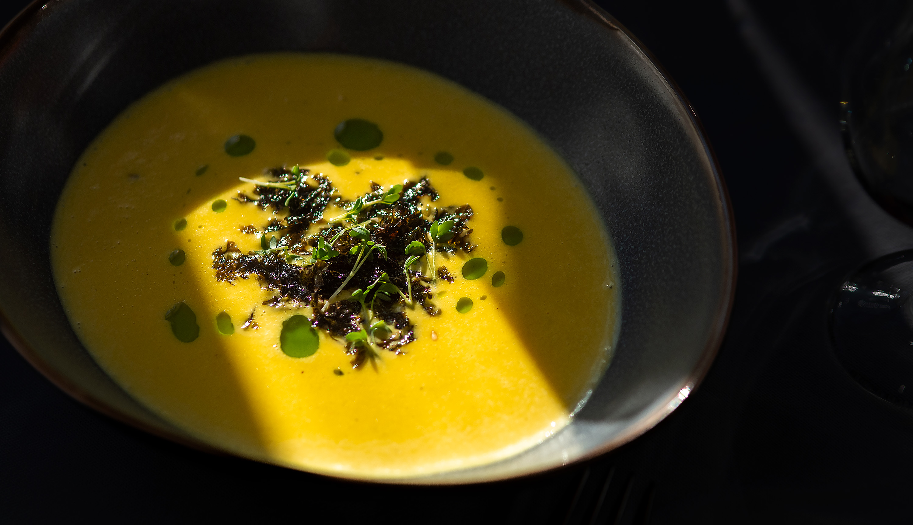 Gul kremet suppe servert i en mørk bolle med grønne urteblad og olje som garnityr.