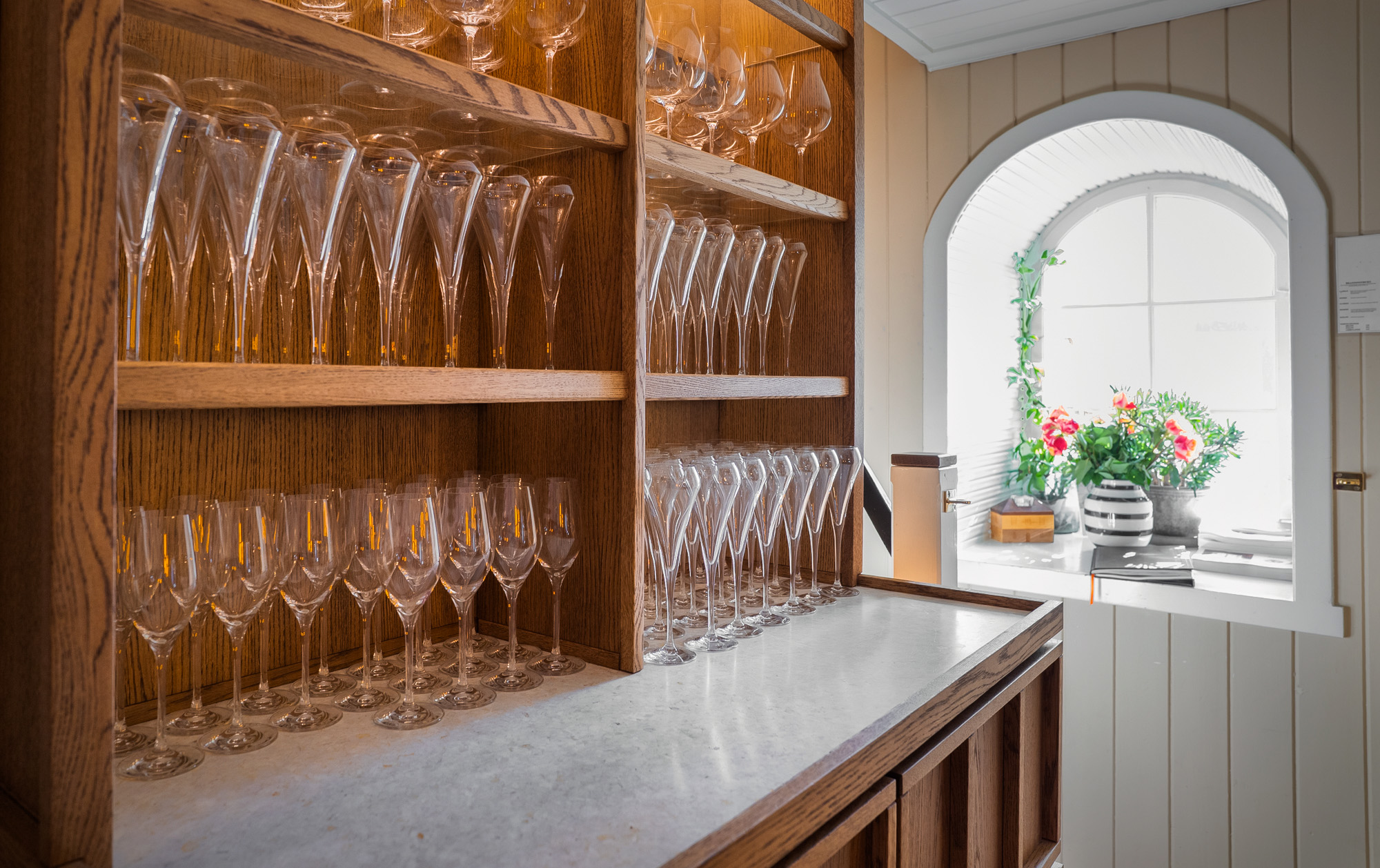 Klar rekke med vinglass av forskjellige størrelser på trehyller ved et vindu med blomster i en stripepott.