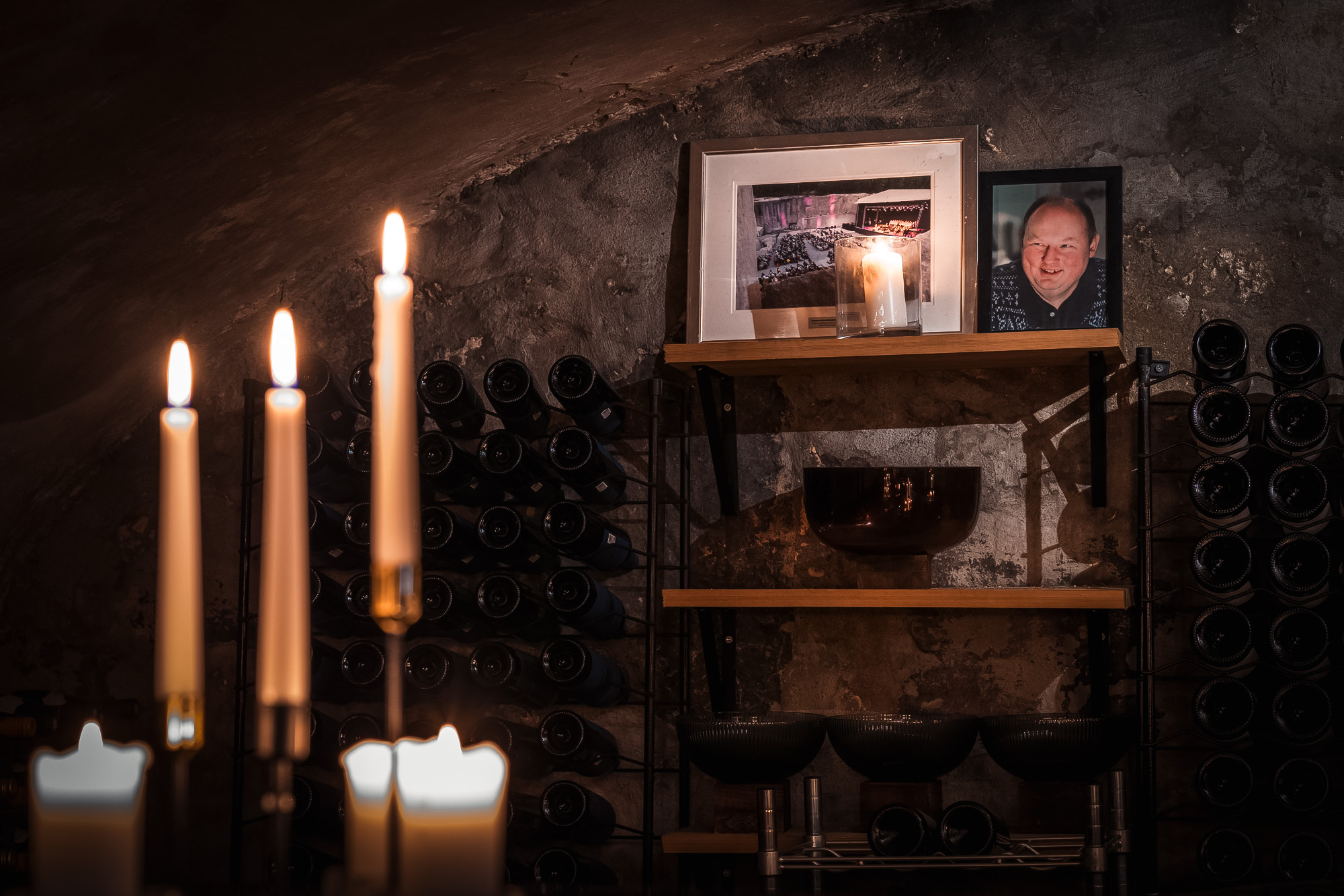 Stearinlys tent i forgrunnen med vinflasker på stativer og tre hylle med bilder og skål i bakgrunnen i et mørkt rom med steinvegg.