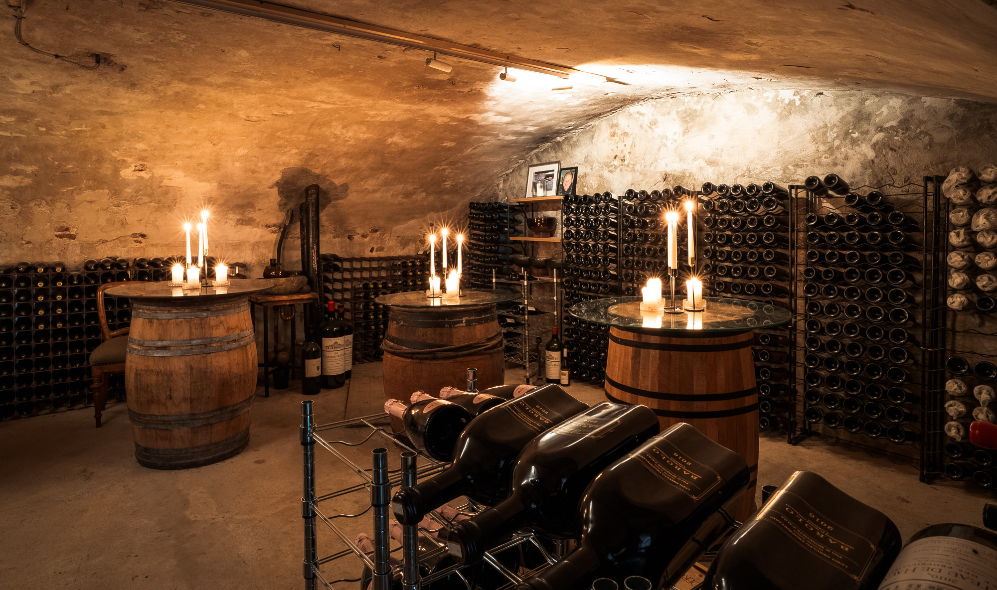 Vinkjeller med tre tønnebord dekorert med levende lys og hyller fylt med vinflasker mot steinvegger.