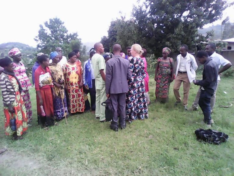 Group of people standing in a circle outdoors on grass, some wearing colorful traditional clothing and one person holding a cane.
