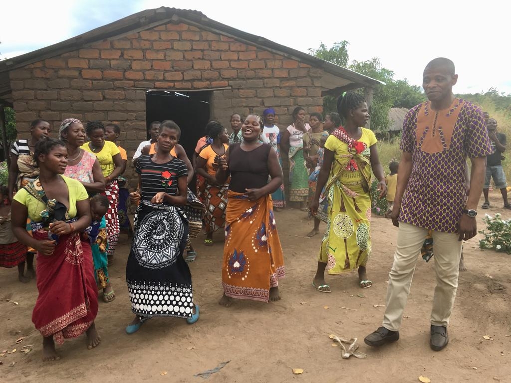 Group of people dressed in colorful traditional clothing gathered outdoors in front of a brick house.