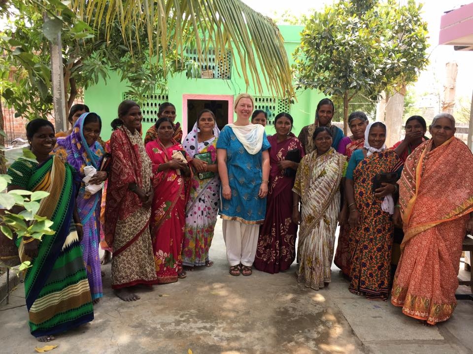 Group of women in colorful traditional Indian sarees standing outdoors in front of a green building, with one woman in blue salwar kameez in the center.