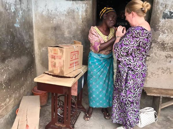 Two women standing and holding hands in a rustic room with a sewing machine table and cardboard box nearby.
