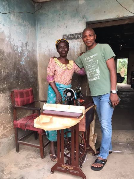 An elderly woman dressed in colorful traditional clothing stands beside a man in a green shirt and jeans next to a vintage sewing machine in a rustic room.