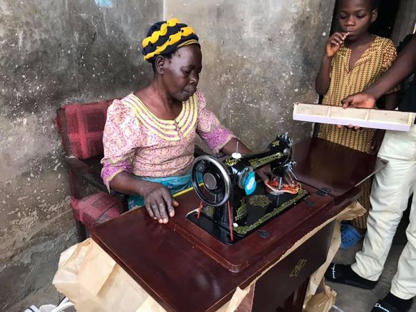 Woman sewing fabric on a vintage black sewing machine at a wooden table, with two children nearby in a rustic room.