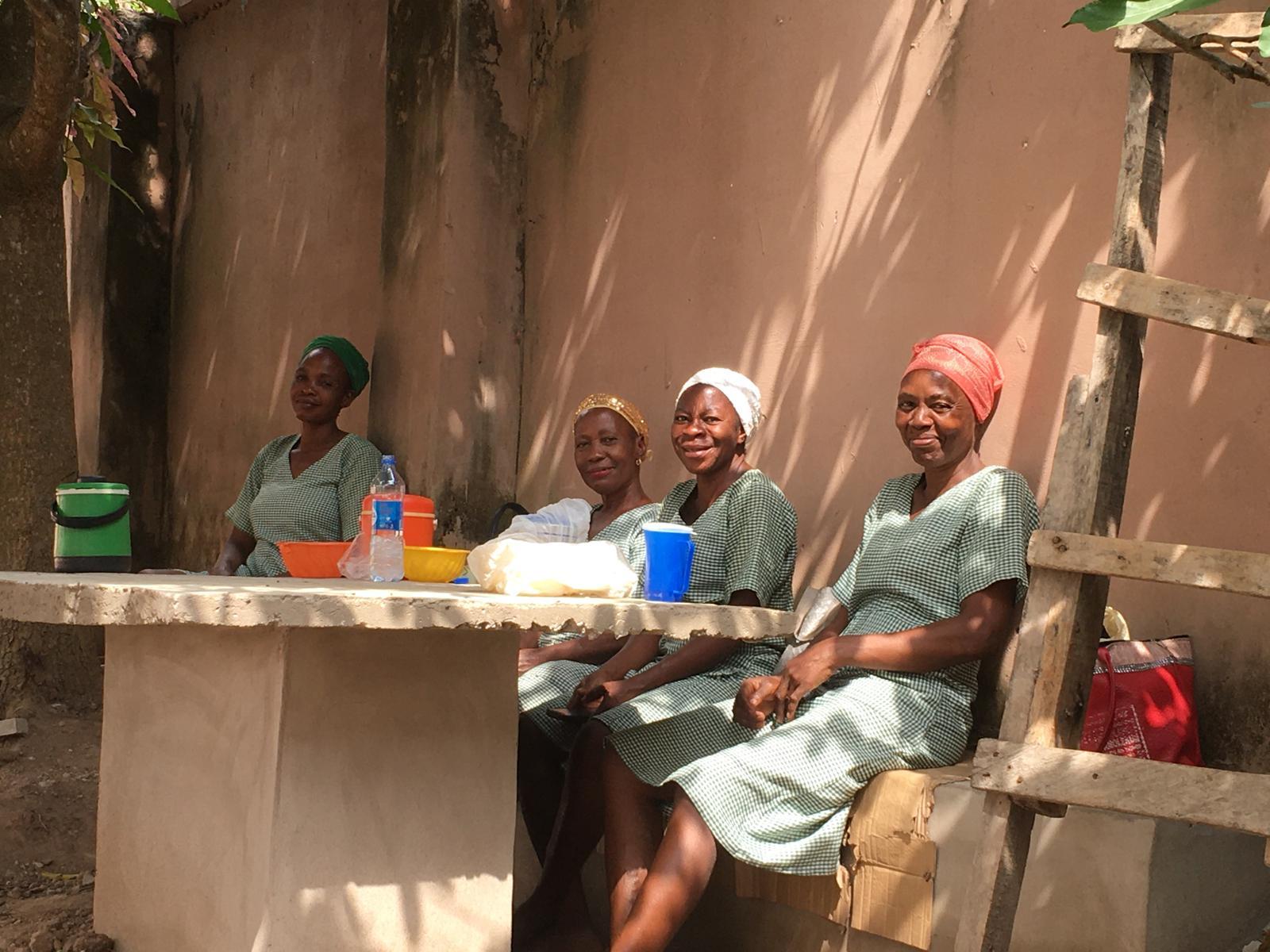 Four women wearing green checkered dresses and headscarves sitting at a concrete table outdoors with food and drink containers.