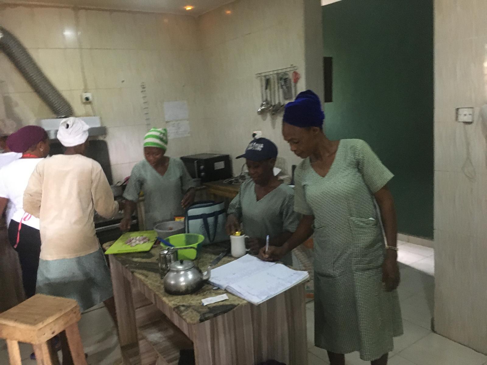 Five women wearing headscarves and uniforms working together in a kitchen around a countertop with cooking utensils and a notebook.