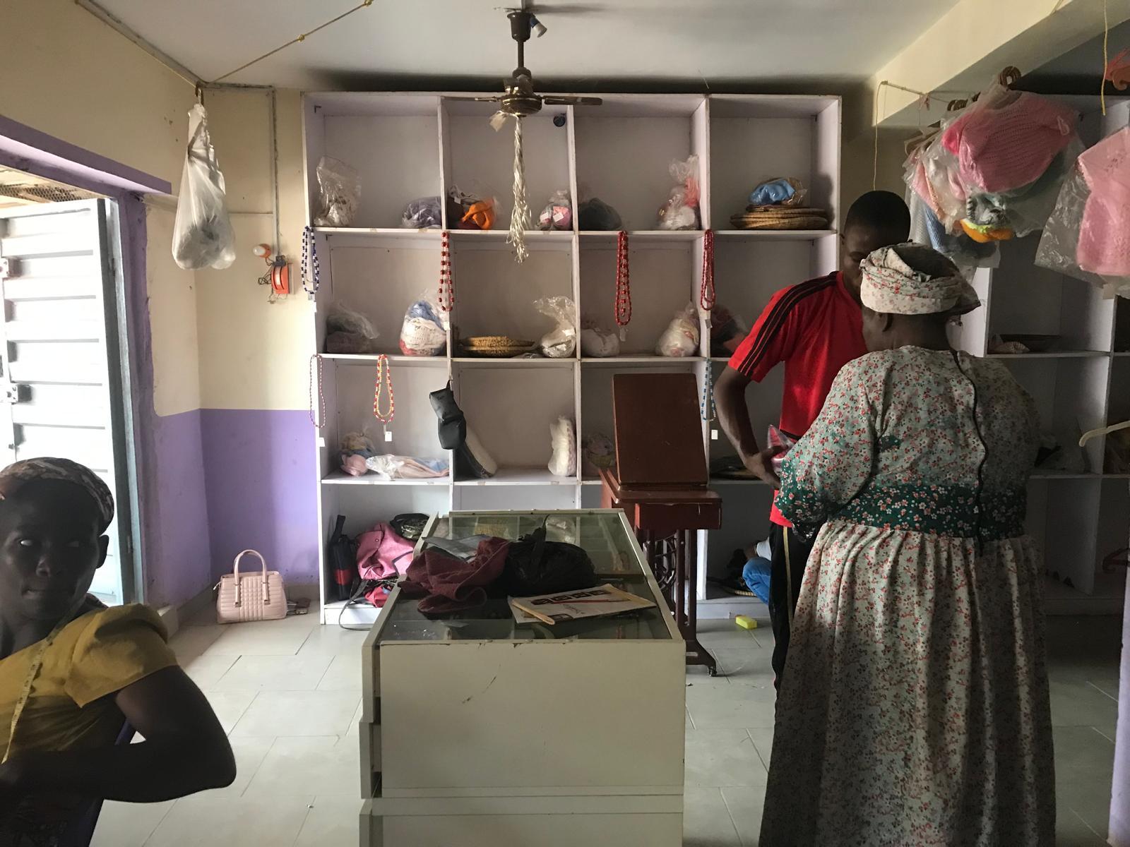 Interior of a small shop with shelves holding various items and two people standing near the counter engaged in conversation.