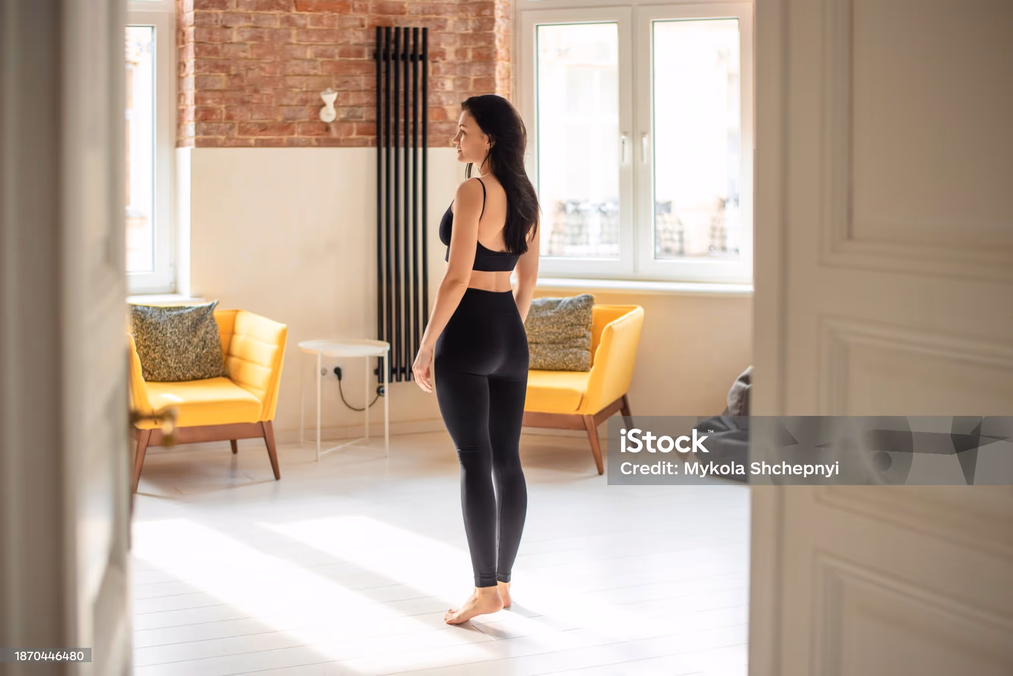 Woman in black workout clothes standing barefoot in a bright room with yellow armchairs and large windows.