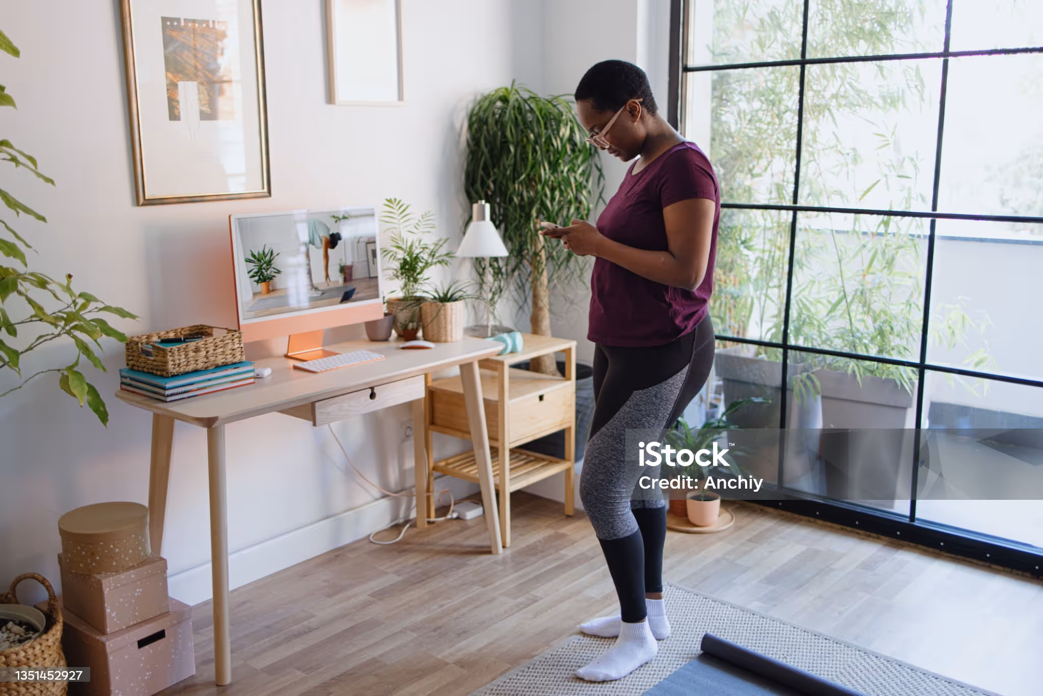 Woman in workout clothes checking her phone in a bright room with plants and a desk showing a yoga video on the computer.