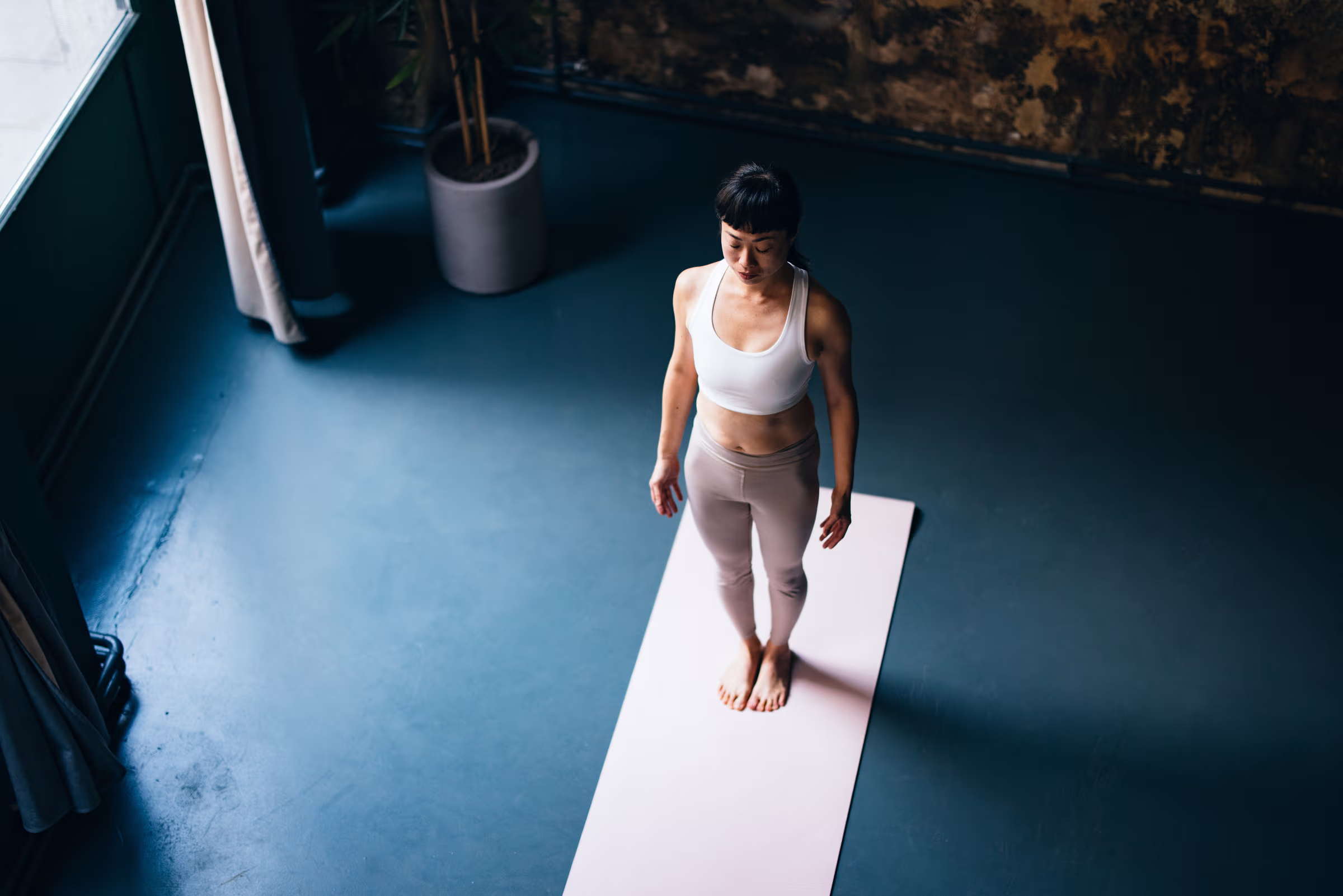 Woman standing quietly on a yoga mat in a dimly lit room with large window and potted plant.