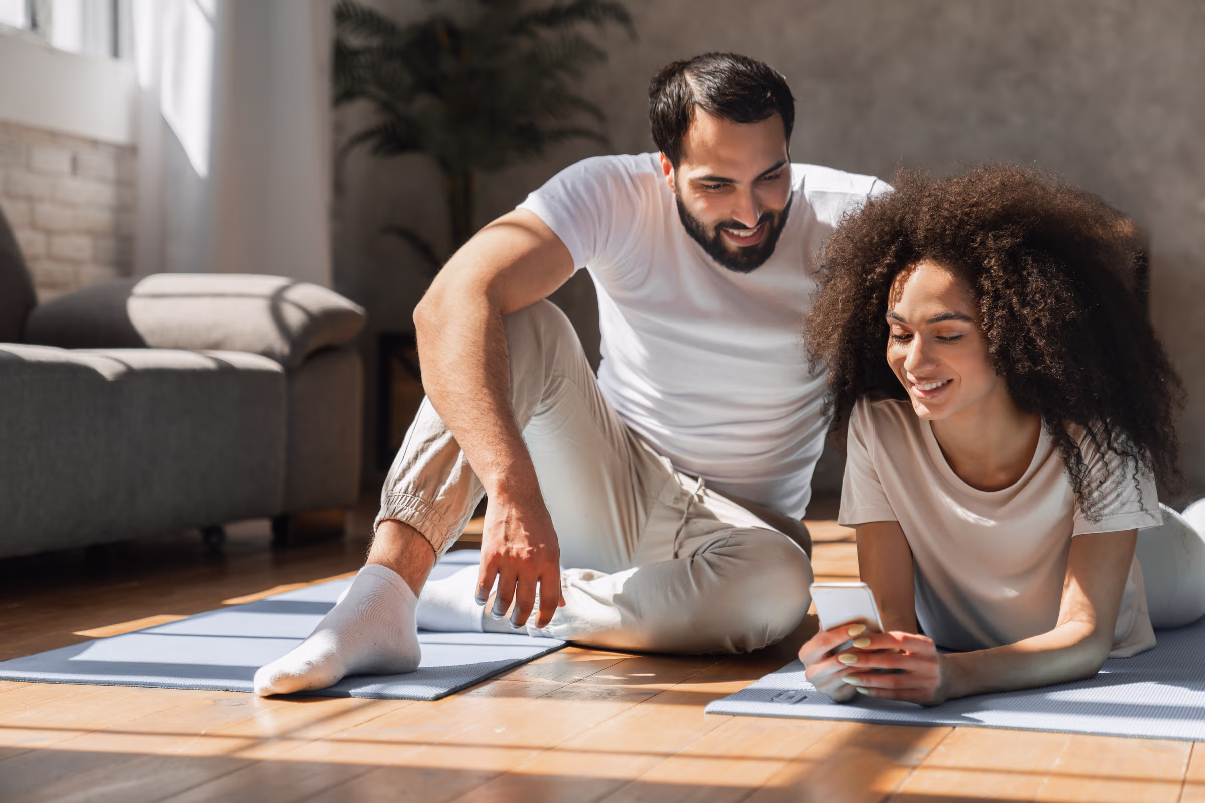 Smiling man and woman in white shirts sitting on yoga mats on a wooden floor, looking at a smartphone together.