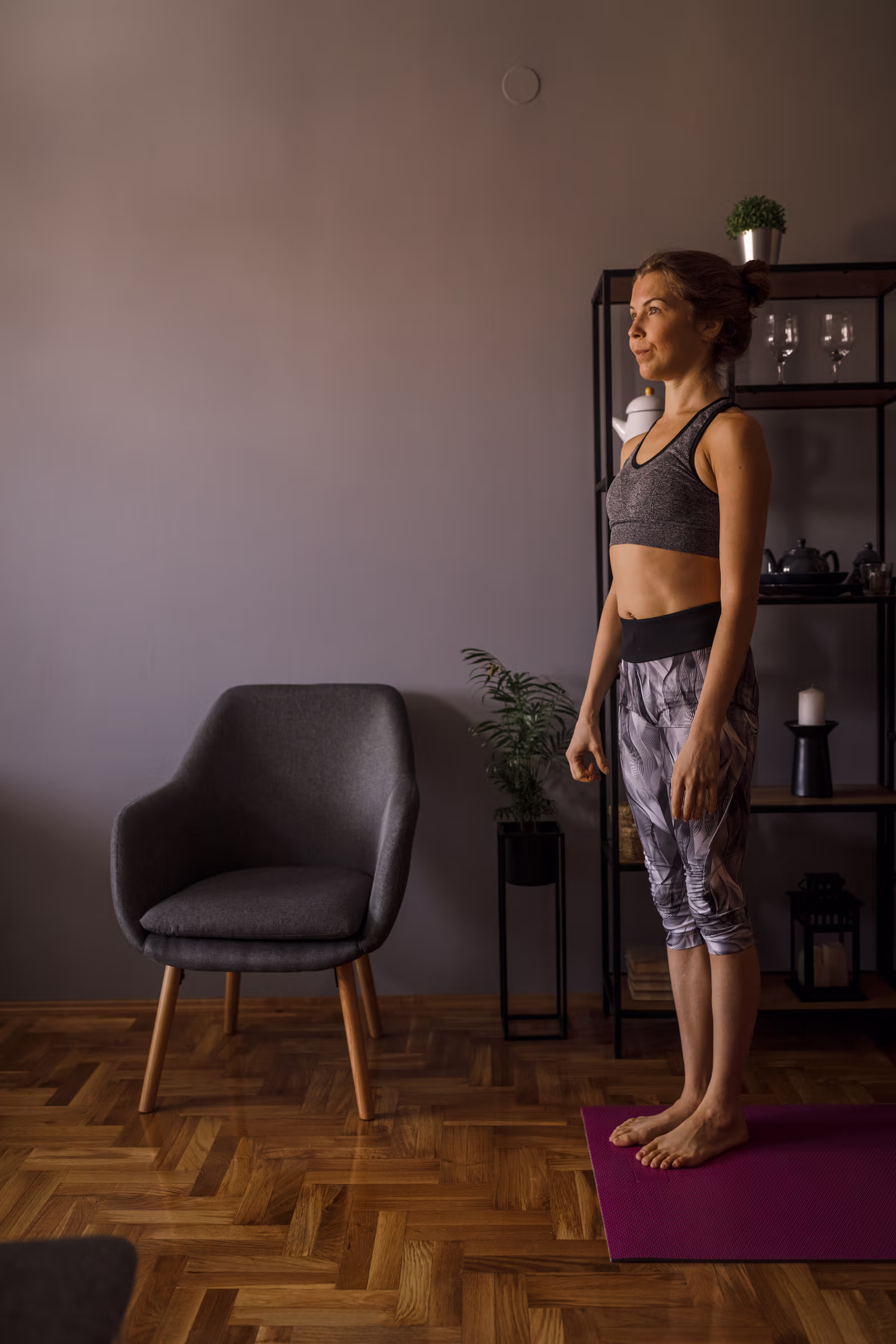 Woman in athletic wear standing on a purple yoga mat in a living room with a gray chair and shelving unit in the background.