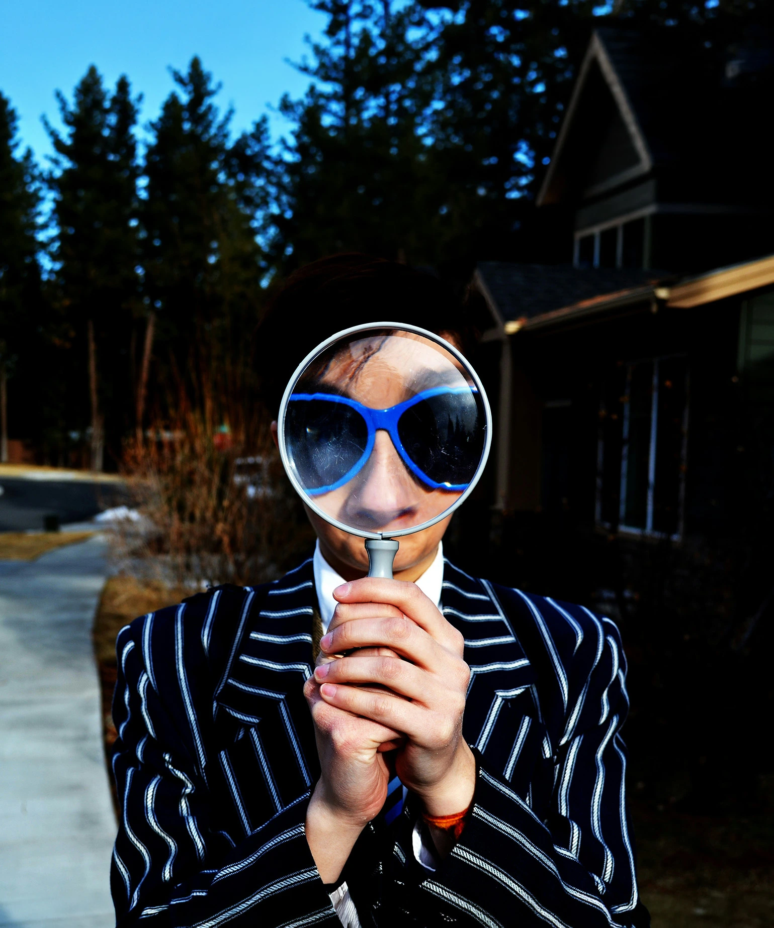 Person in a striped blazer holds a magnifying glass in front of their face, enlarging blue sunglasses, while standing on a sunlit path with trees and houses in the background.