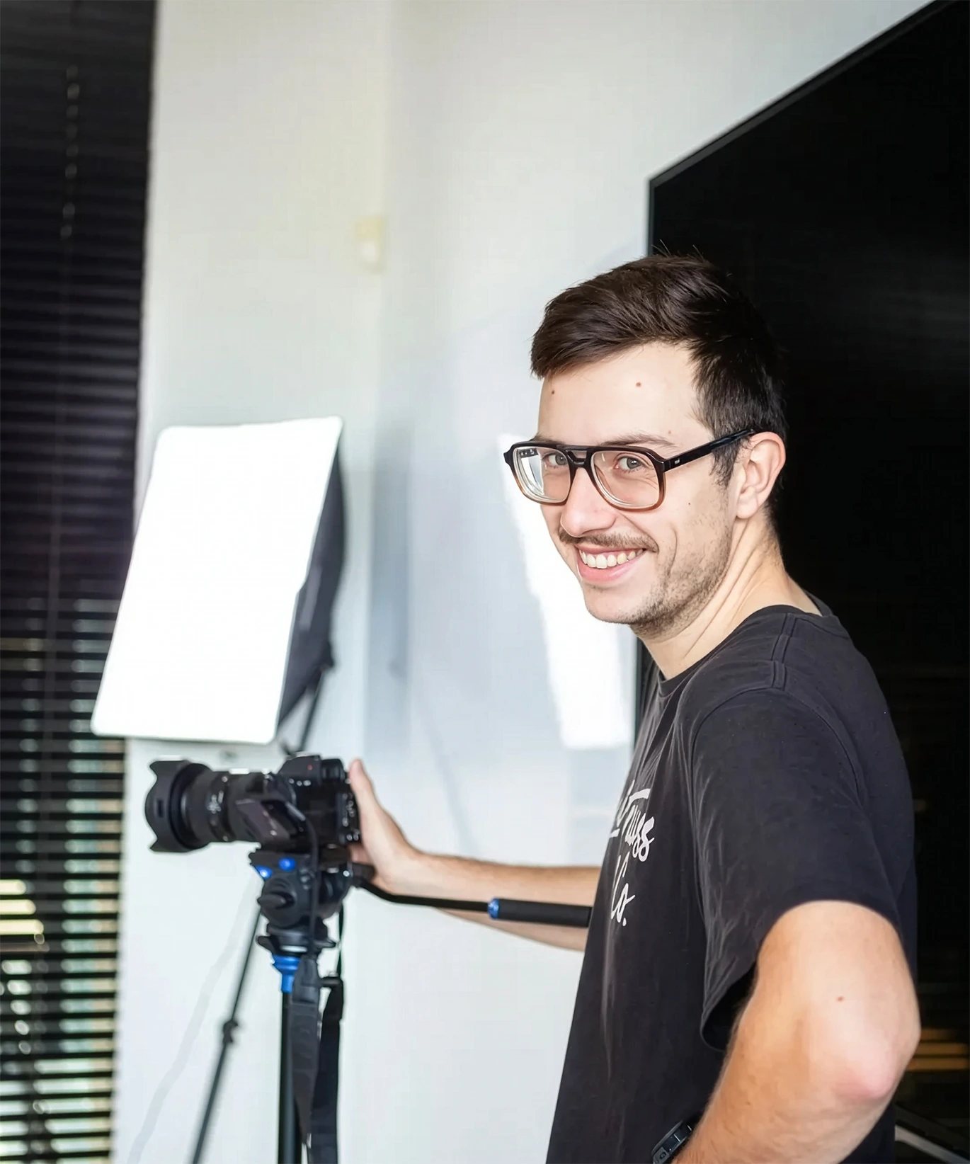 Smiling man wearing glasses stands beside a camera on a tripod with a bright studio light, in a modern indoor workspace.
