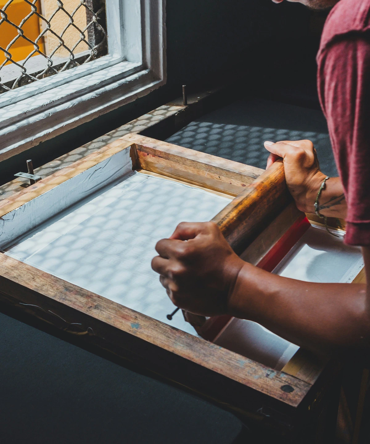 Person using a wooden screen-printing frame and squeegee to spread ink across fabric on a worktable near a window with patterned light.