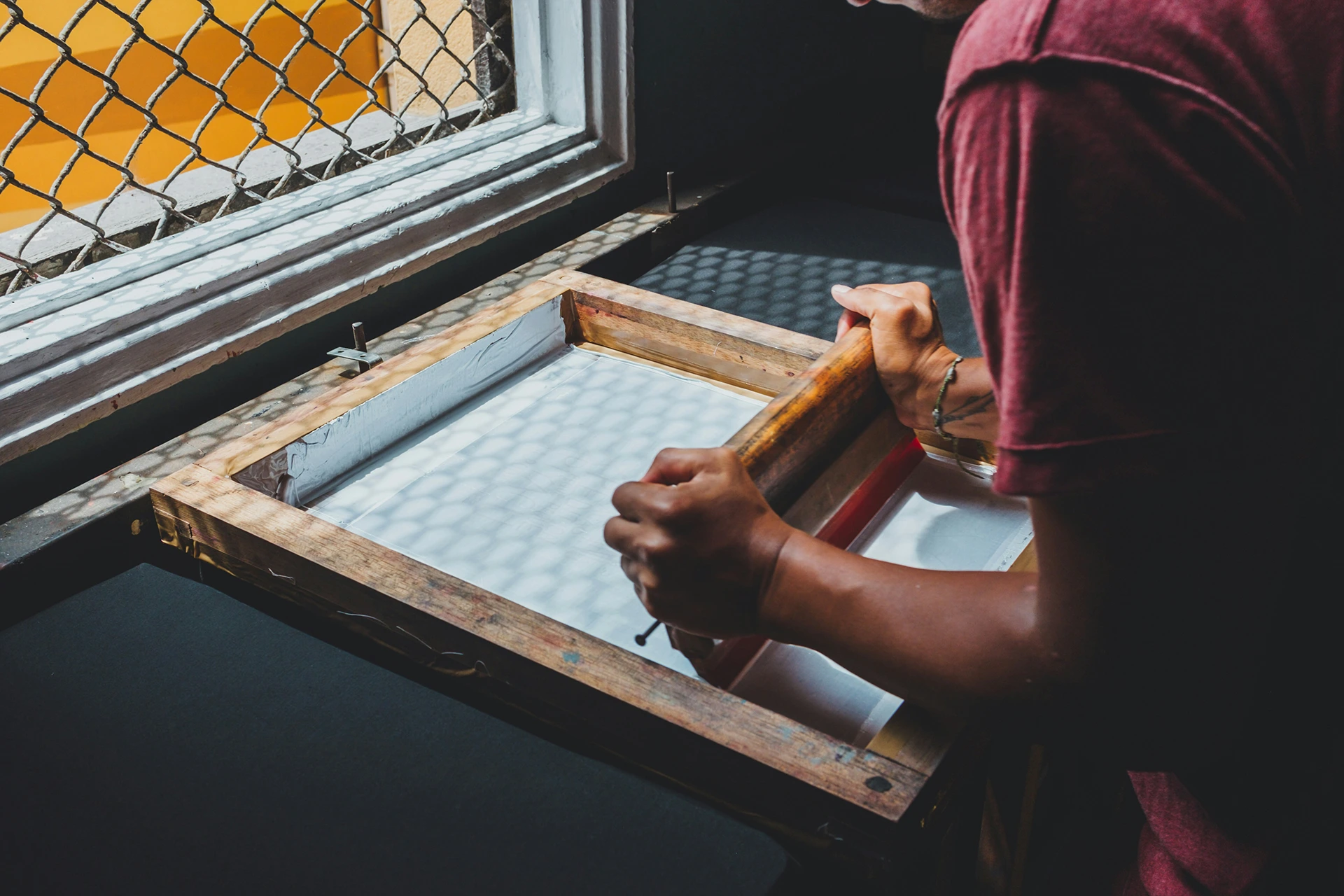 Person using a wooden screen-printing frame and squeegee to spread ink across fabric on a worktable near a window with patterned light.