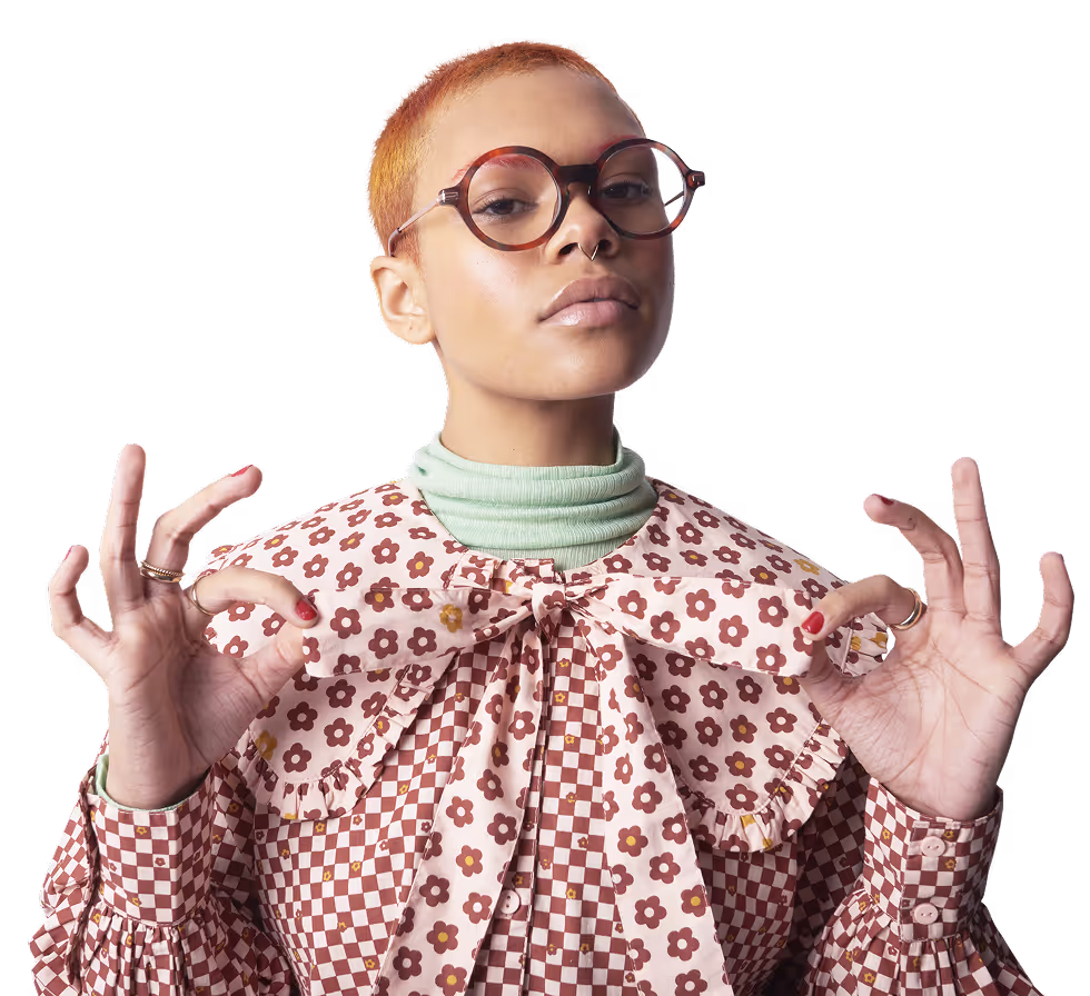 Jeune femme aux cheveux courts teints en orange, portant de grandes lunettes rondes et une chemise à motifs floraux et à carreaux, faisant un geste OK avec les deux mains.