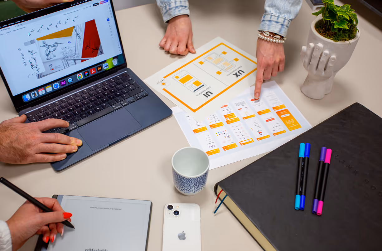 Trois personnes travaillant ensemble sur la conception UX, avec un ordinateur portable affichant des dessins, des schémas imprimés, une tablette graphique, un smartphone et un carnet de notes sur la table.