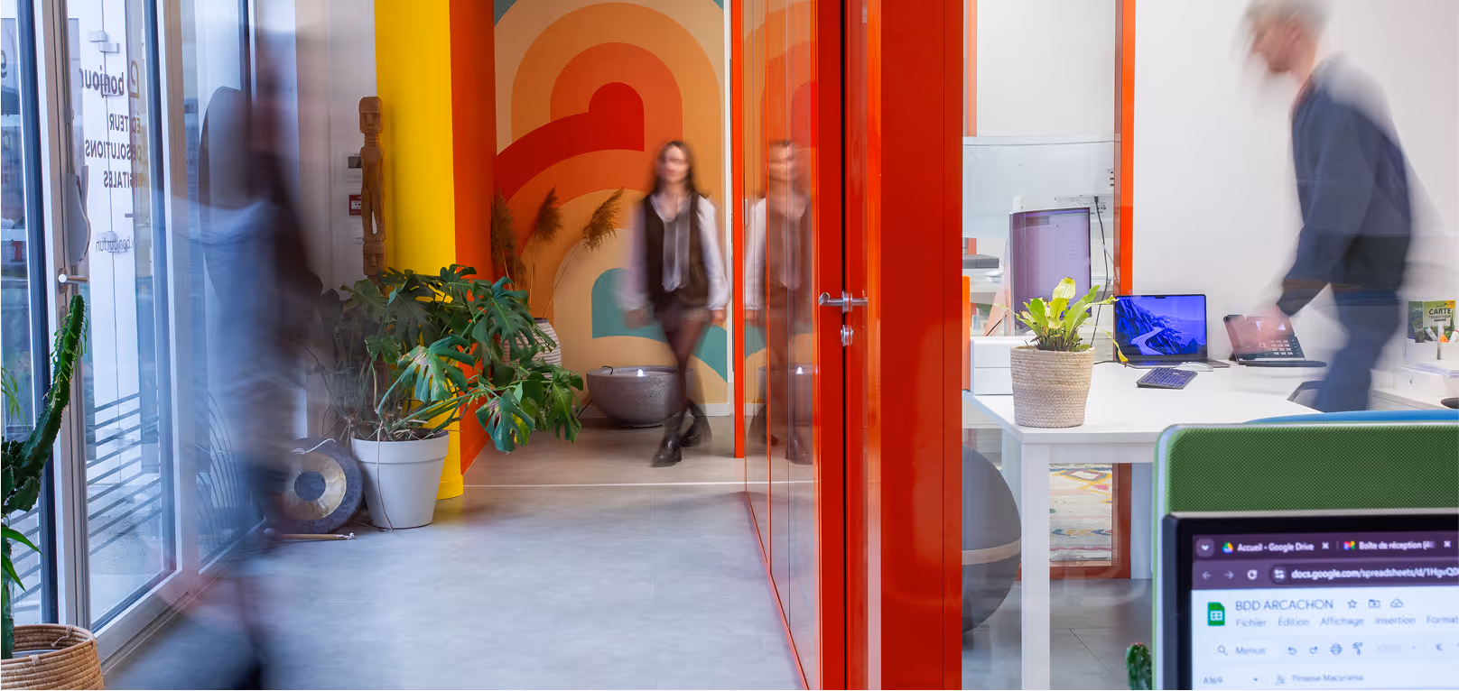 Bureau moderne avec cloisons en verre rouge, plantes vertes, décor coloré et personnes en mouvement flou.