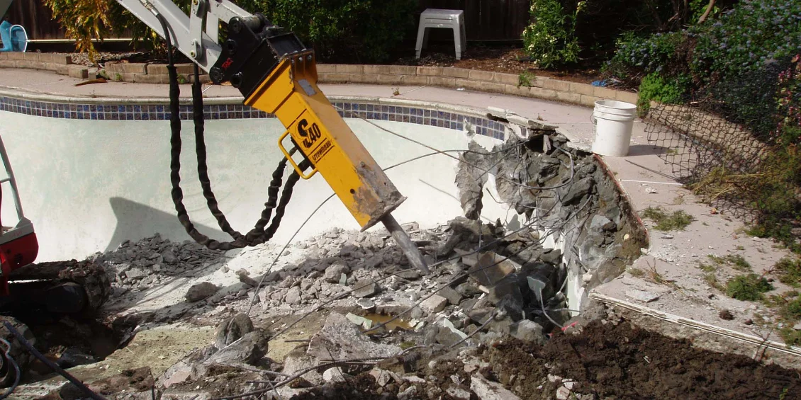 Heavy excavator machinery performing pool demolition by breaking concrete walls, illustrating the labor-intensive pool removal cost factors.