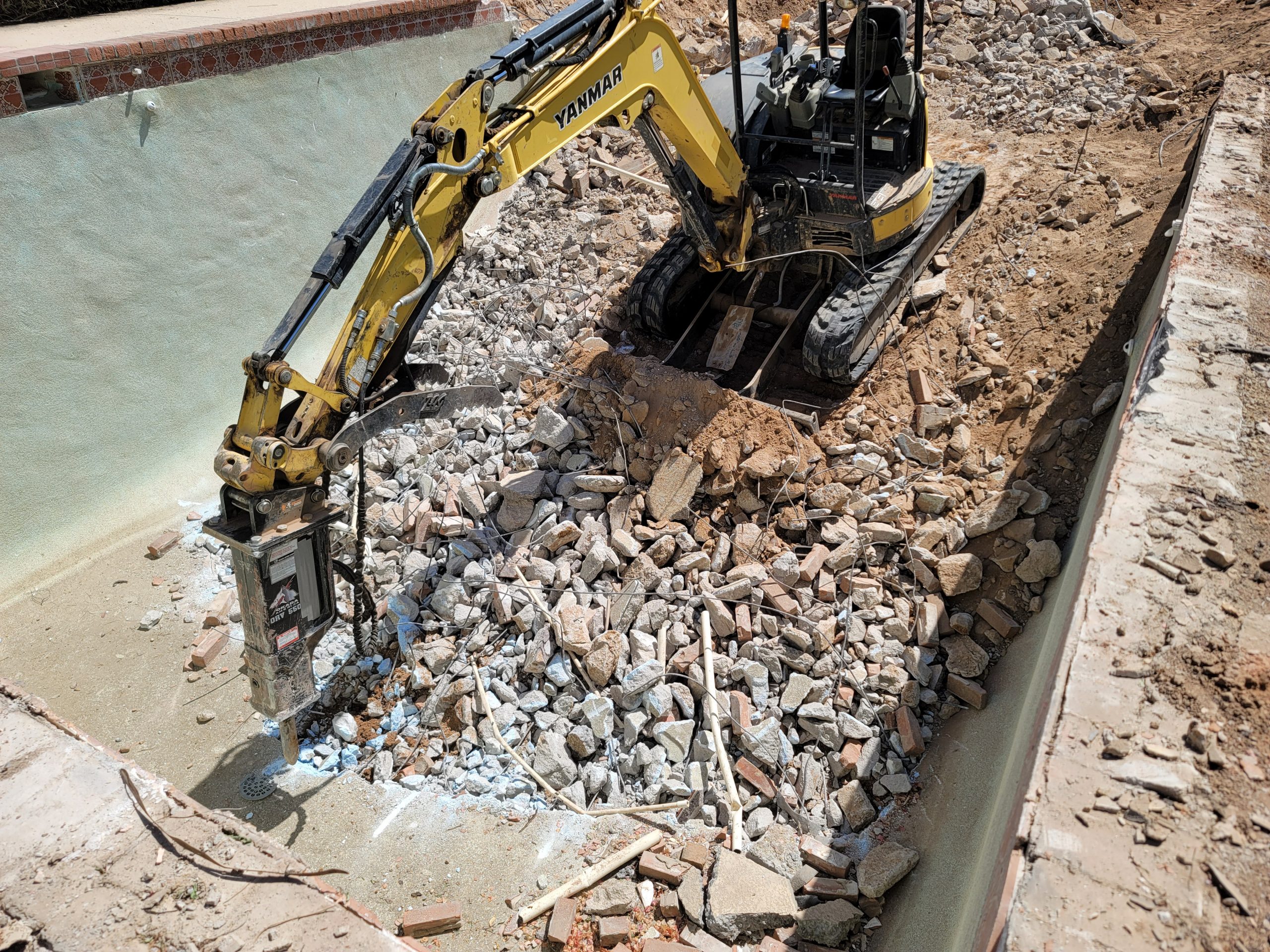 A miniature excavator rental unit with retractable tracks squeezing through a narrow townhouse garden gate to begin swimming pool demolition.