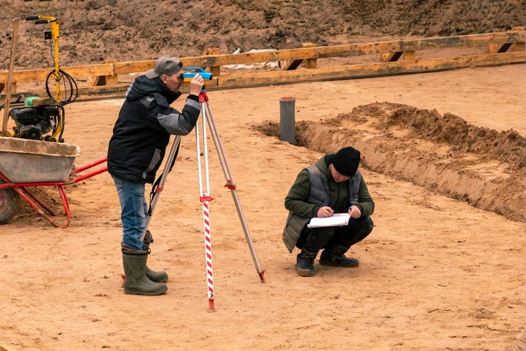 A surveyor using a theodolite/laser level on a construction site to measure the height difference between the road curb and the plot soil.