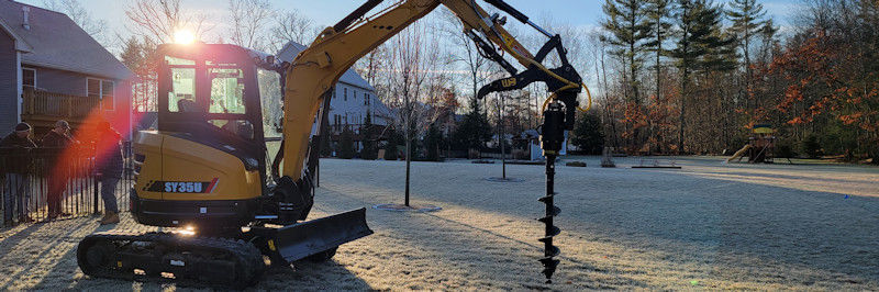 A Bobcat excavator auger attachment drilling deep holes near a sidewalk, demonstrating versatility for both fencing and digging holes for trees.