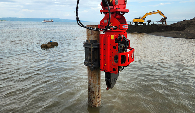 Close-up of a Side-Grip vibro hammer clamping onto a sheet pile lying on the ground, demonstrating the self-loading capability.