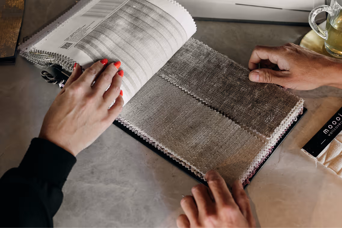 Twee handen bladeren door een stoffencollectie met neutrale beige en grijstinten op een tafel.