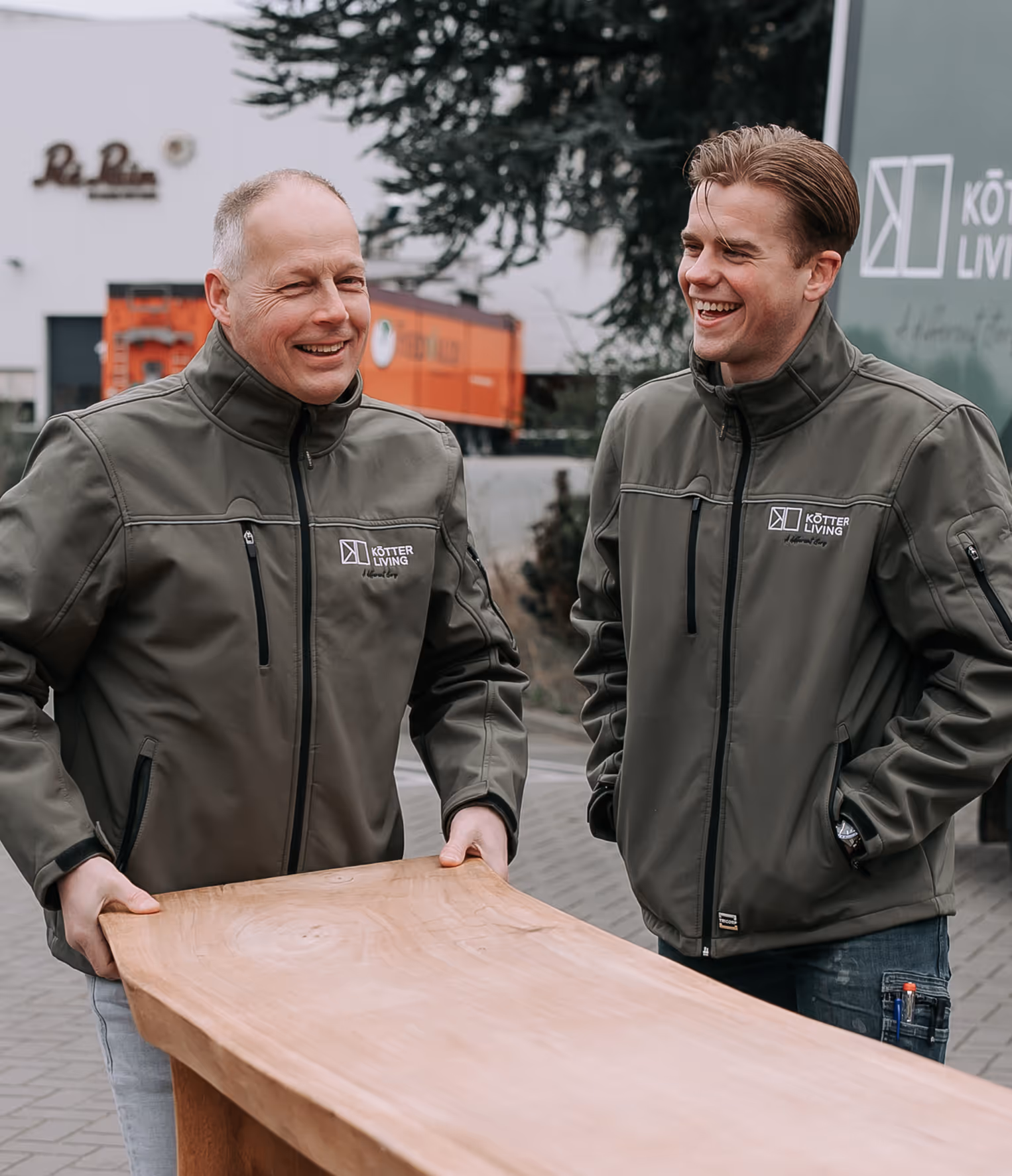 Twee lachende mannen in grijze jassen met 'Kötter Living' logo tillen samen een houten meubelstuk buiten.