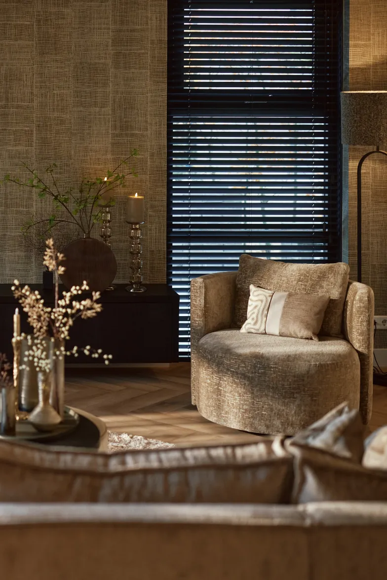 Gezellige woonkamerhoek met een ronde beige fauteuil, kussens, een zwarte vensterbank met brandende kaarsen en een staande lamp naast een raam met donkere jaloezieën.