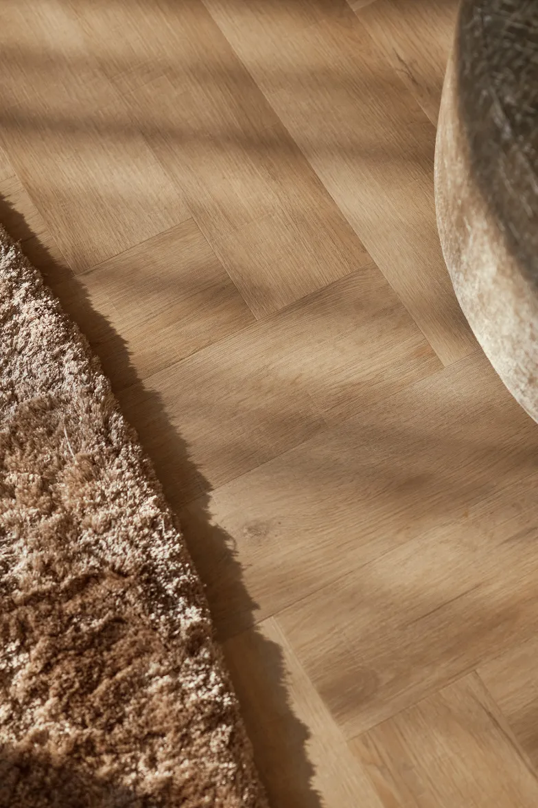Lichtbruin houten visgraatvloer met beige pluizig tapijt en deel van een ronde beige stoel.