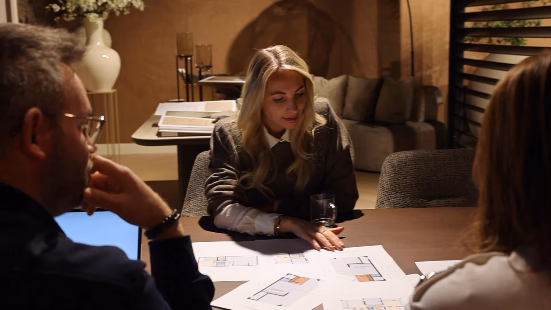 Drie mensen bespreken plattegronden aan een tafel in een warme, gezellige ruimte met een vaas en een bank op de achtergrond.