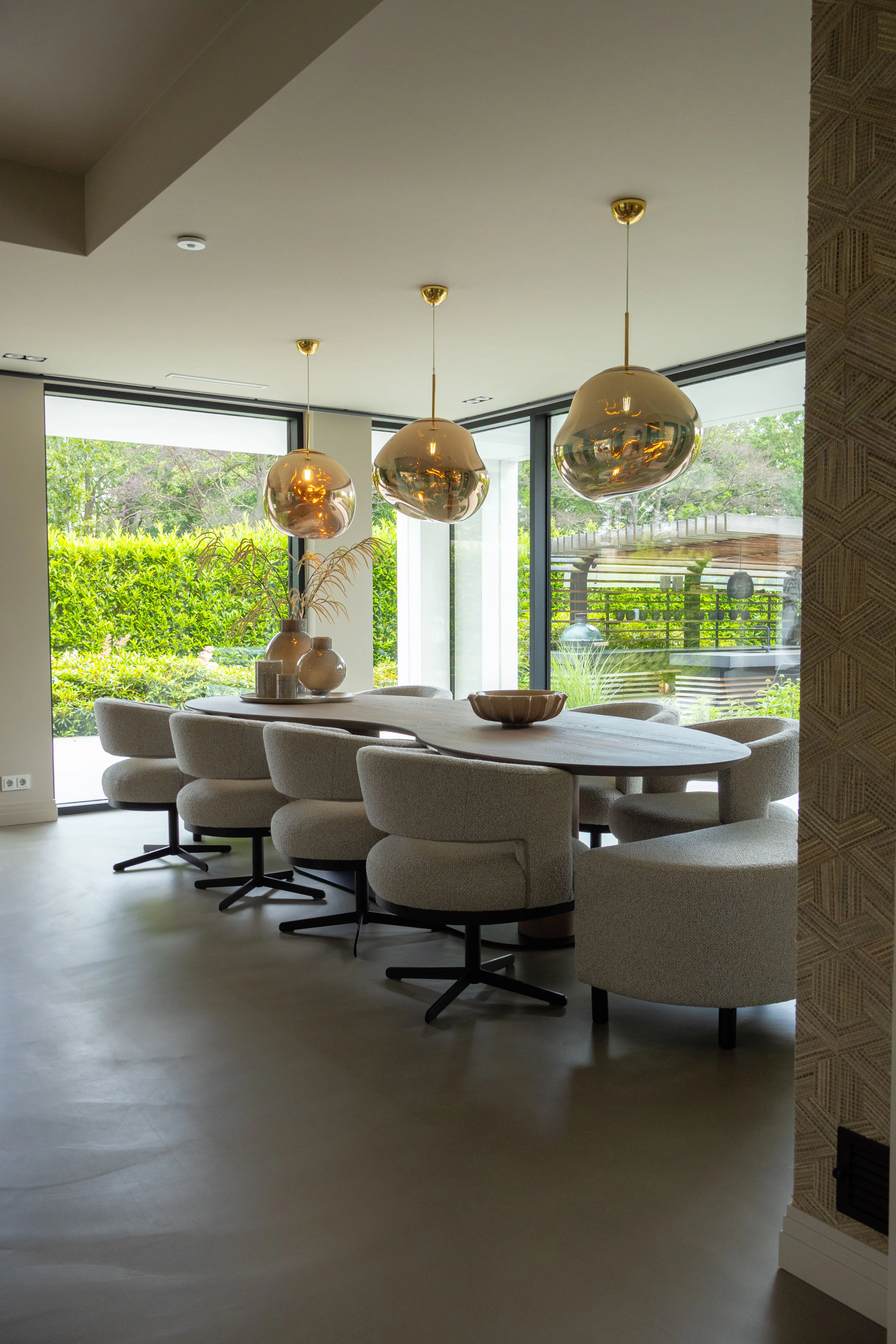 Moderne keuken met een organisch gevormde eettafel, stijlvolle eetkamerstoelen en grote hanglampen boven de tafel. Grote ramen zorgen voor veel natuurlijk daglicht.
