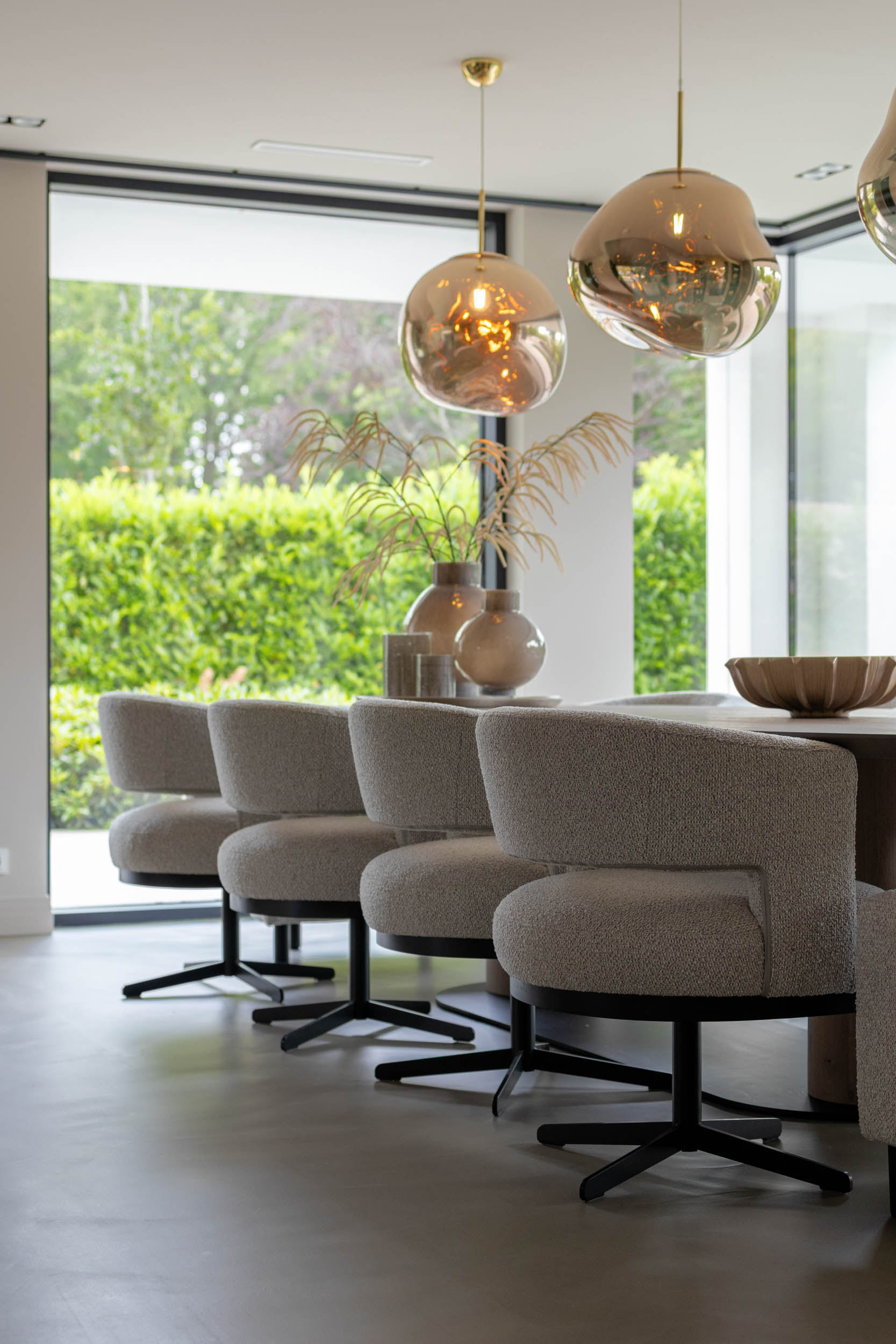 Moderne keuken met een organisch gevormde eettafel, stijlvolle eetkamerstoelen en grote hanglampen boven de tafel. Grote ramen zorgen voor veel natuurlijk daglicht.