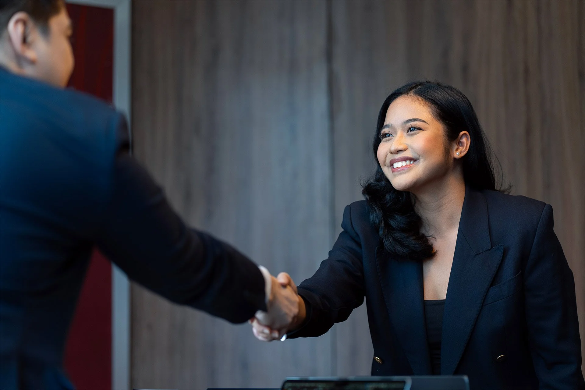Smiling woman in a dark blazer shaking hands with another person during a professional meeting.
