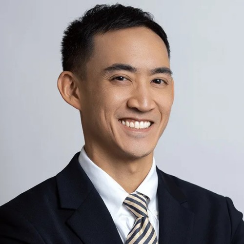 Smiling man in a dark suit, white shirt, and striped tie against a light gray background.