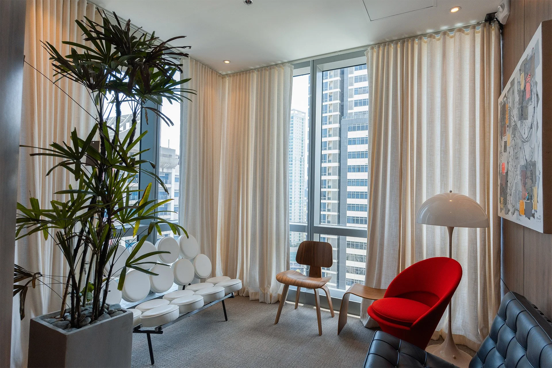 Modern office lounge with large windows, cream curtains, a tall plant in a gray pot, white circular-padded bench, wooden chair, red curved chair, and white floor lamp.